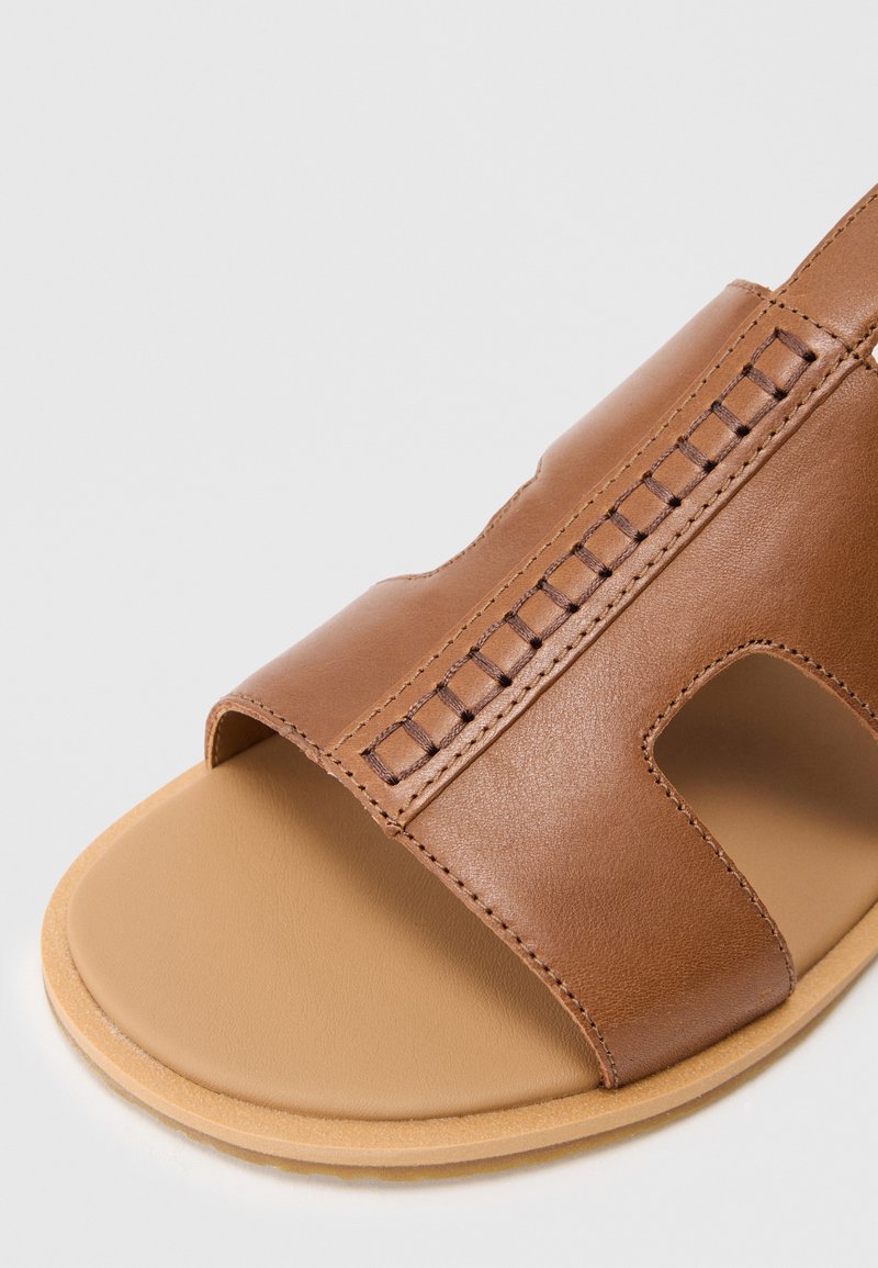 Bruine leren sandaal met open teen, brede banden en decoratieve stiksels. Zachte tan kleurige zool met een gladde textuur. Minimalistisch ontwerp.