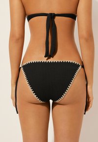 Czarny dół bikini z teksturowanej tkaniny, z wiązanym bocznym krojem i kontrastującym falbanym wykończeniem na brzegach.