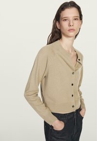 Cardigan in lana beige, con collo tondo, bottoni neri e maniche lunghe. Tessuto morbido con un taglio corto e leggera costina all'orlo.