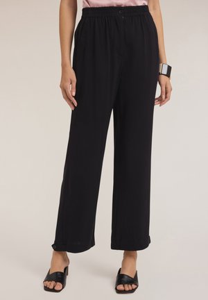 Zwarte wijde broek met een elastische tailleband en een knoop aan de voorkant. Heeft zijzakken en licht opgerolde zoom. Gecombineerd met zwarte instap sandalen.