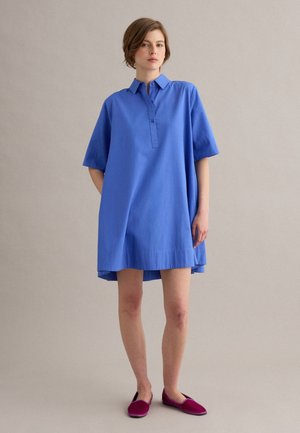 Joven mujer de pie, vestida con un vestido camisero suelto azul de manga corta y zapatos planos magenta, frente a un fondo neutro y liso.