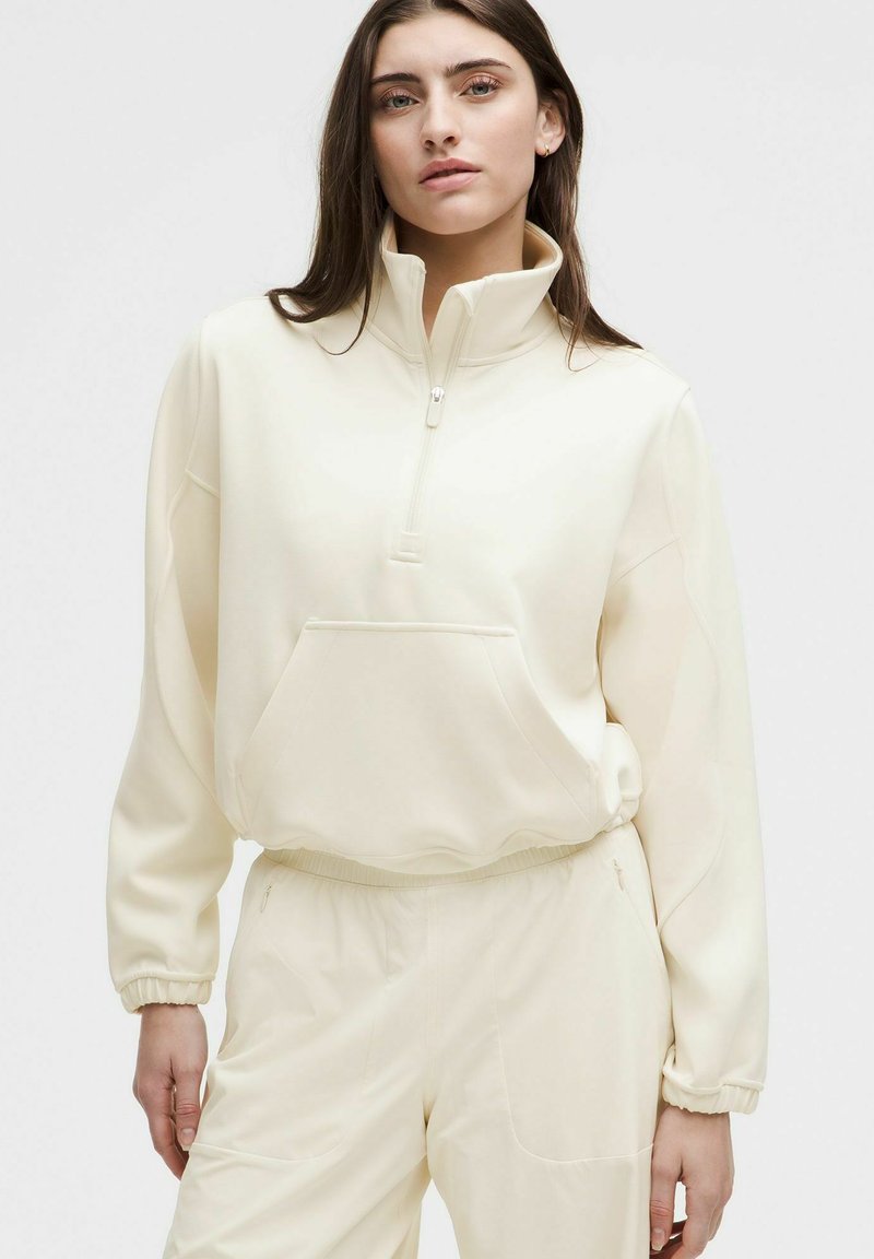 Sudadera blanca con cremallera, cuello alto, corte holgado, bolsillo delantero y puños fruncidos, confeccionada en tela suave y texturizada.