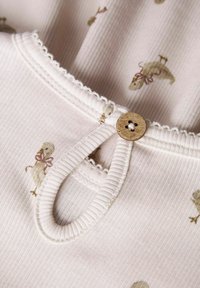 Tela color crema con textura acanalada, que presenta un estampado de dinosaurios y un botón de madera en el cuello. Costuras decorativas en ondas en los bordes.