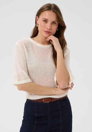 Femme aux longs cheveux bruns portant un pull blanc à manches courtes et une jupe bleu foncé avec une ceinture marron, posant avec la main près du visage.