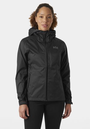 Mujer con chaqueta impermeable negra con capucha, cremallera frontal y bolsillos laterales con cremallera, de pie frente a un fondo claro y liso.