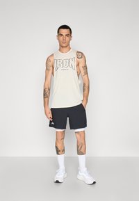 Men's sleeveless beige tank top with "IRON" graphic in black, paired with dark shorts. White socks and sneakers complete the athletic look.