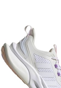 Zapatilla deportiva blanca con un upper texturizado, paneles de malla y suela de goma. Presenta acentos morados y una lengüeta en el talón.