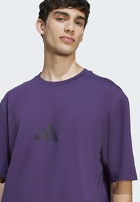 Violet trumpas rankovėmis marškinėliai, pagaminti iš minkštos medžiagos, su juodu adidas logotipu krūtinėje ir apvaliu kaklu.