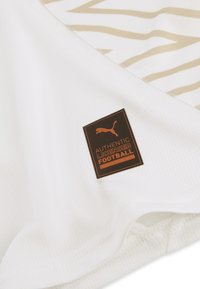 Puma fodboldtrøje i hvidt stof med mesh-struktur, med et guldstribet design og et brunt logo-patch, der står "AUTHENTIC LICENSED FOOTBALL."