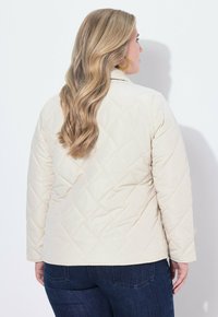 Frau mit langen blonden Haaren, die goldene Creolen, eine cremefarbene gesteppte Jacke und dunkelblaue Jeans trägt, mit dem Rücken zur Kamera.