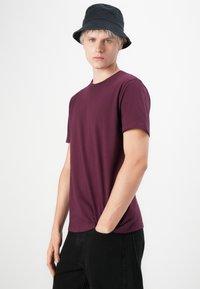 Burgundi színű, rövid ujjú póló sima, könnyű anyagból, fekete nadrággal és sötétkék vödörkalappal párosítva. Minimalista design.