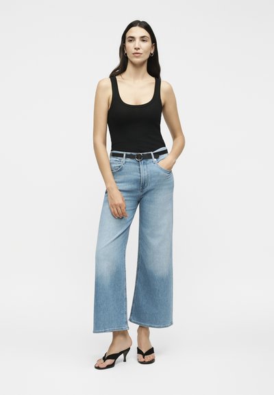 Γυναίκα που φοράει μαύρο τοπ χωρίς μανίκια, ανοιχτό μπλε τζιν με φαρδιά παντελόνια και μαύρη ζώνη, και μαύρα σανδάλια με τακούνι, στέκεται με το ένα χέρι στην τσέπη.
