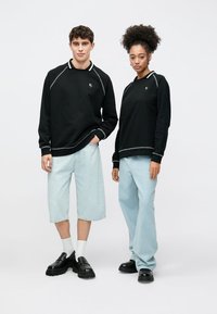 Μαύρο φούτερ με λευκά ράμματα, άνετη εφαρμογή· συνδυασμένο με ανοιχτό μπλε τζιν σορτς και ανοιχτό μπλε τζιν παντελόνι. Μαύρα παπούτσια ολοκληρώνουν την εμφάνιση.