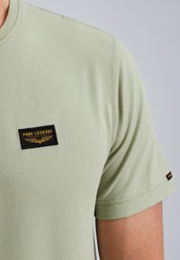 Svetlozelené bavlnené tričko s okrúhlym výstrihom, krátkymi rukávmi a čiernym obdĺžnikovým nášivkom s množstvom zlatého textu a krídlami.