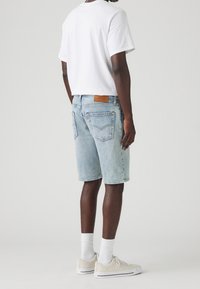 Muž v světle modrých denimových šortkách, bílém tričku, bílých ponožkách a krémových teniskách, stojící na pozadí čistě bílé stěny.