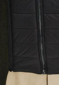 Czarna ocieplana kurtka z zamkiem błyskawicznym o średniej długości, z pikowaną strukturą. Z jednej strony przyszyta do ciemnozielonego materiału.