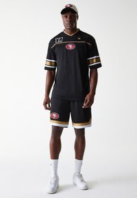 Svart sportsdrakt med gullige detaljer og logo, kombinert med matchende svarte shorts med hvit kant, båret med en baseballcaps.