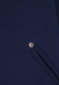 Marinblått tyg med slät textur, med en silverknapp med logotyp och en sidoficka.