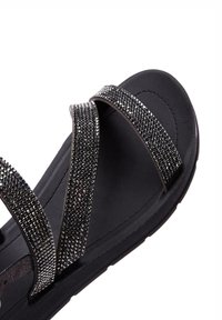 Sandale noire avec des brides croisées ornées d'embellissements métalliques. La semelle est plate avec une surface texturée pour une meilleure adhérence.