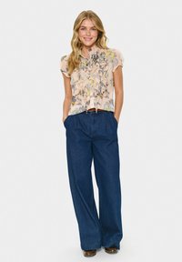 Smiling woman with long blonde hair wearing a floral ruffled blouse, wide-leg dark blue jeans, and brown shoes, standing with hands in pockets.