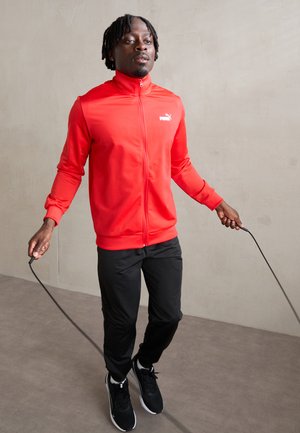 Red zip-up athletic jacket with a white logo, paired with black pants and black sneakers. The model holds a jump rope in a neutral background.
