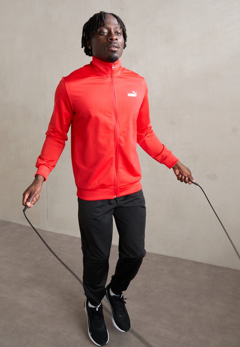 Rode zip-up sportjack met een wit logo, gecombineerd met zwarte broek en zwarte sneakers. Het model houdt een springtouw vast op een neutrale achtergrond.