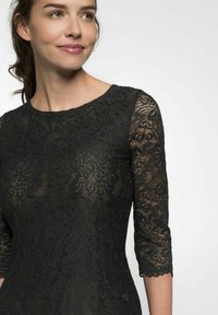 Schwarzes Spitzenkleid mit 3/4-Ärmeln, rundem Ausschnitt, floralen Mustern und gewellten Kanten. Besitzt einen Knopfakzent an der Taille.