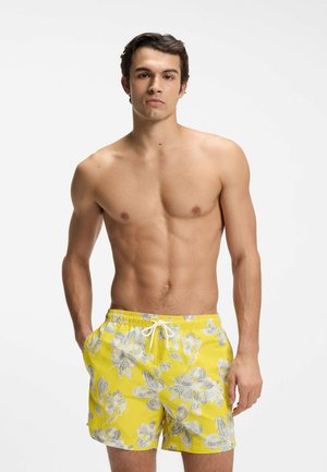 Junger Mann mit kurzen dunklen Haaren, der gelbe geblümte Badeshorts mit weißem Kordelzug trägt, steht mit den Händen in den Taschen vor weißem Hintergrund.