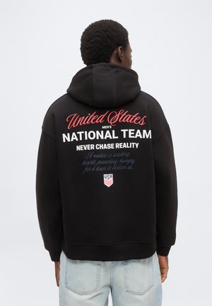 Mężczyzna o krótkich kręconych włosach, ubrany w czarną bluzę z kapturem z napisem „United States Men's National Team” oraz motywacyjnym tekstem na plecach, jasnoniebieskie dżinsy.