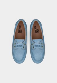 Ζευγάρι ανοιχτόμπλε μοκασίνια από σουέντ με χρυσές οπές για κορδόνια και την επωνυμία "Love Moschino" στις εσωτερικές σόλες και τις γλώσσες τους, σε λευκό φόντο.