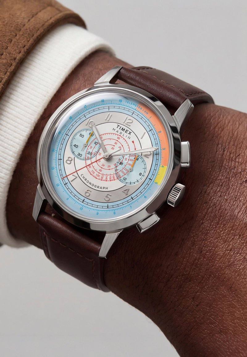 Хронографски часовник Timex Marlin с бял циферблат, многоцветни маркери, сребърен корпус и кафява кожена каишка, носен на китка с тъмна кожа.