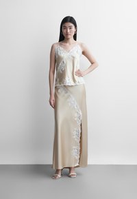 Žena stojící opřená o bílou zeď, má na sobě krémové saténové tílko a odpovídající dlouhou sukni s bílými krajkovými detaily a sandály s otevřenou špičkou.