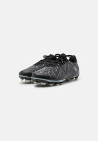 Botas de fútbol negras con una parte superior sintética lisa, diseño en zigzag gris, textura perforada y clavos moldeados sobre una suela blanca.