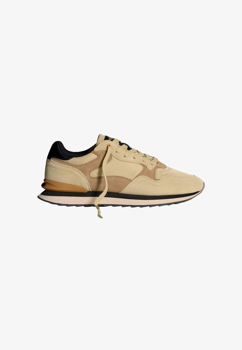 Sneaker in suede beige con accenti neri, design con lacci, suola ammortizzata e sovrapposizioni testurizzate. Presenta una linguetta sul tallone di colore marrone in contrasto.