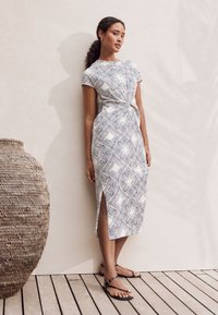 Witte jurk met zwart geometrisch bladvormig patroon, korte mouwen en zijsplitten; gecombineerd met zwarte platte sandalen. Geweven mand op de voorgrond.