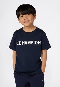 Tamno plava pamukasta majica s bijelim logom "CHAMPION" sprijeda. Kratki rukavi i okrugli izrez, nošena s odgovarajućim tamnoplavim hlačama.