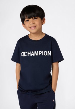 Temno modra bombažna majica s kratkimi rokavi in zaobljenim izrezom, na sprednji strani z belim logotipom "CHAMPION". Nosena z usklajenimi temno modrimi hlačami.