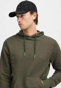 Olivově zelená mikina s textured látkou, nastavitelné stahovací kapuce, přední klokanka kapsa a světle zelené kovové doplňky. Černá čepice.