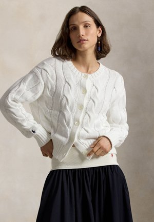 Γυναίκα που φοράει λευκή ζακέτα με πλεχτό σχέδιο και κουμπιά, μπεζ ζώνη μέσης, μπλε φούστα και κρεμαστά σκουλαρίκια, ποζάροντας μπροστά σε απλό φόντο.