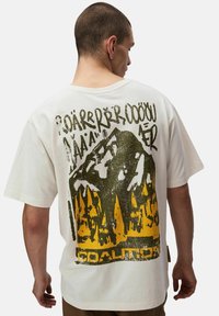 Mężczyzna w białej koszulce z graficznym motywem góry i lasu w czerni i żółci oraz napisem "KOALICJA" na dole.