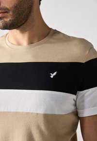 Beige a černé pruhované tričko vyrobené z měkké bavlny, které má malé bílé vyšívané logo ptáčka na levé straně hrudi. Kroužkový výstřih.