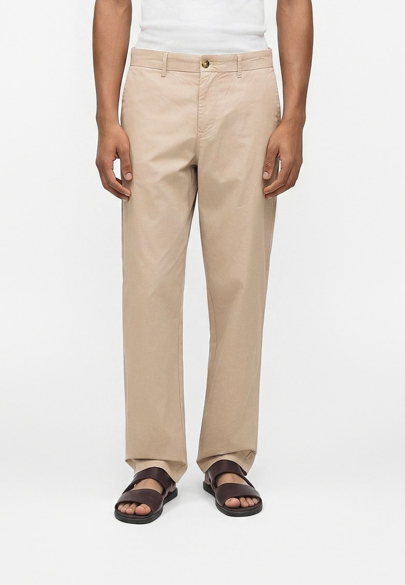 Άνδρας που φοράει μπεζ παντελόνια ίσιας γραμμής, λευκό τοπ και σκούρα καφέ ανοιχτά σανδάλια, στέκεται σε λευκό φόντο.