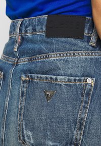 Jeans in denim con un lavaggio blu sbiadito, caratterizzati da un'etichetta in pelle nera con il logo "GUESS 81" in rilievo e un logo metallico triangolare sulla tasca posteriore.