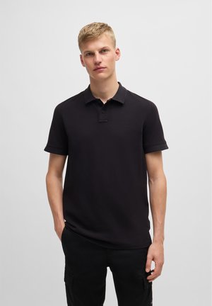 Polo negro hecho de algodón con una tapeta de dos botones, mangas cortas y un sutil logo en el pecho. Llevado con pantalones negros.
