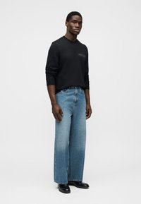 Μαύρο φούτερ με στρογγυλή λαιμόκοψη και λεπτομέρεια με λογότυπο, συνδυασμένο με ανοιχτό μπλε τζιν παντελόνι φαρδύ στο κάτω μέρος. Το μοντέλο φοράει μαύρα μποτάκια. Απλός, μίνιμαλ σχεδιασμός.