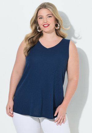 Femme souriante aux longs cheveux blonds porte de grandes boucles d'oreilles argentées, un haut bleu marine sans manches et un pantalon blanc sur un fond blanc.