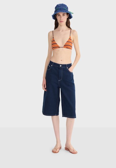 Femme portant un short long en denim bleu, un haut de bikini rayé orange et marron, un chapeau bob bleu, et des tongs beige, debout devant un fond uni.