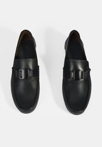 Zwarte leren loafers met een verstelbare gesp, een gladde textuur, een ronde neus en contrasterende stiksels op een flexibele zool.