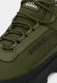 Grønn sneakers laget av teksturert materiale, med runde snører, en diskret Sorel-logo og en svart gummisåle. Akenter i en lysere grønn.