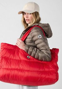 Dikke bruine jas met capuchon, rode oversized schoudertas en lichtbeige baseballcap. De tas heeft een logo en een gewatteerde textuur.